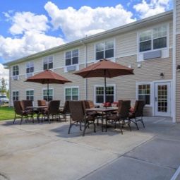 An outdoor patio area with tables, chairs, and umbrellas set up in front of a two-story building under a partly cloudy sky.
