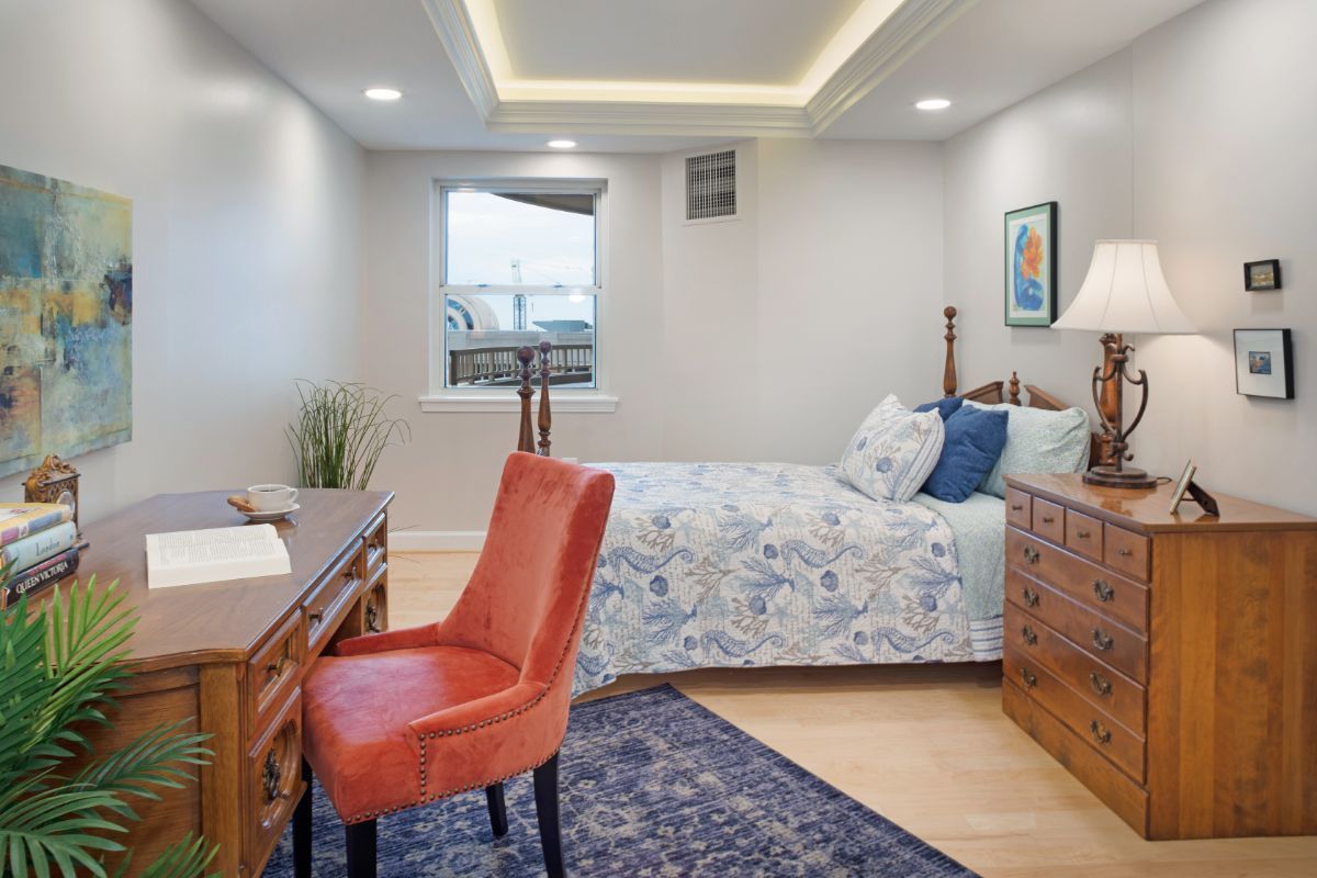 A cozy bedroom featuring a wooden desk and chair, a neatly made bed with floral bedding, and soft lighting.