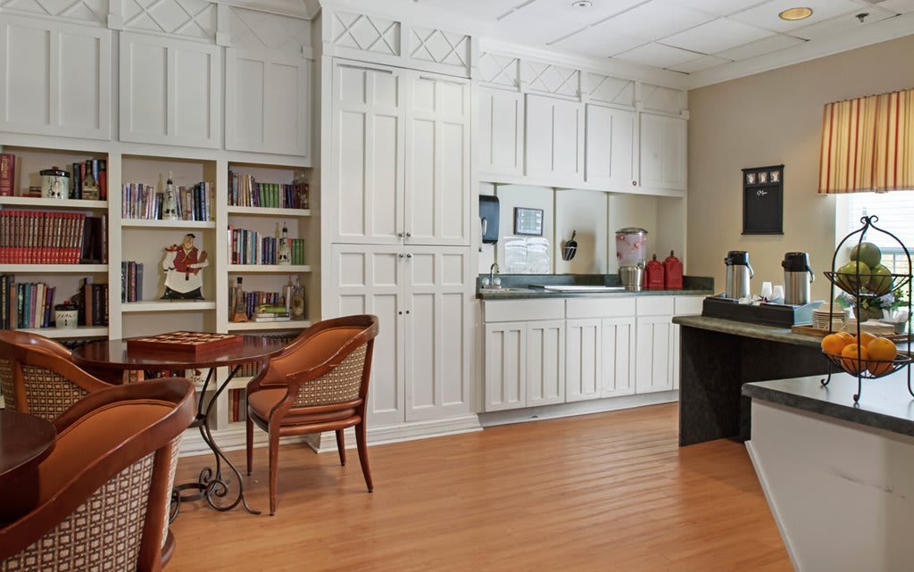 A bright kitchen area with white cabinets and an integrated workspace, featuring a wooden desk and chair alongside a built-in bookshelf.