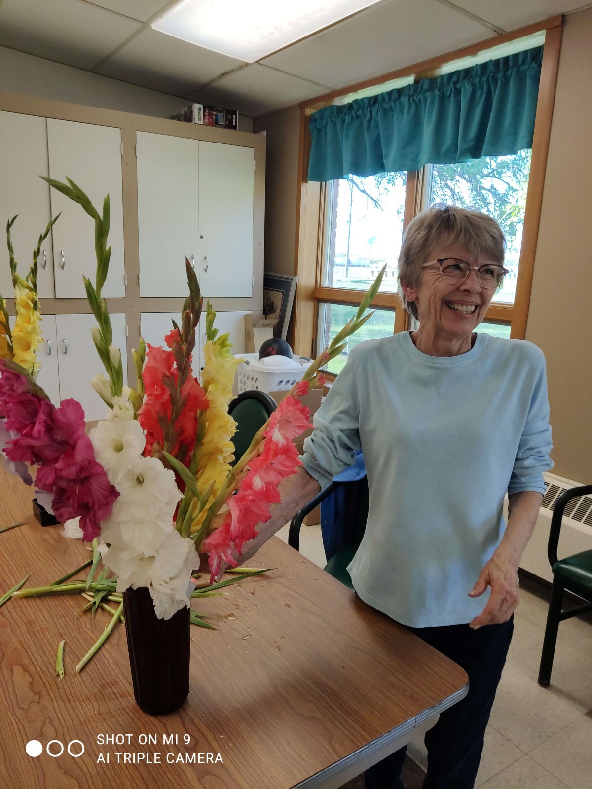 A woman smiling indoors next to a colorful flower arrangement on a table.