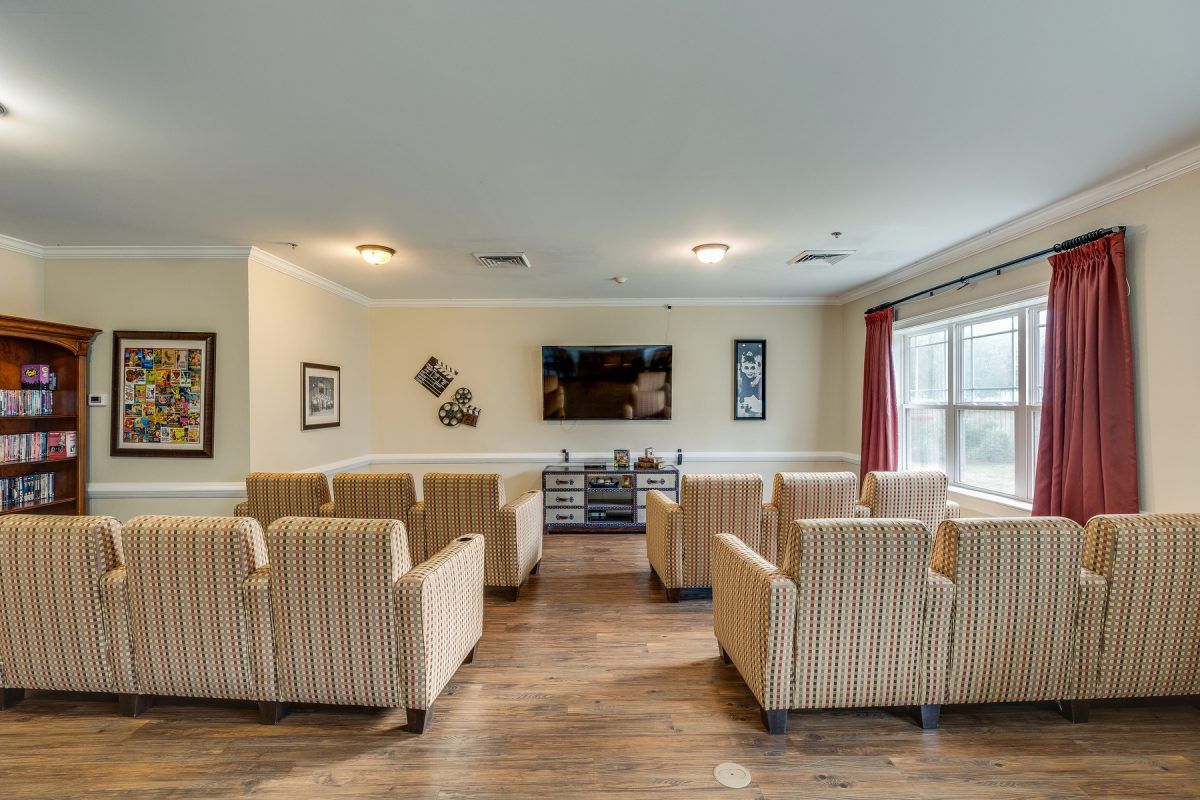 A cozy seating area with multiple chairs facing a television, featuring wooden flooring and large windows.