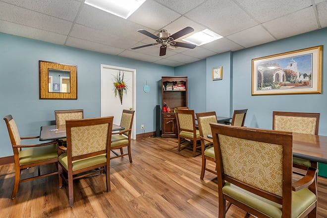 A cozy dining area with wooden flooring, featuring several tables and chairs, ceiling fan, and blue-painted walls adorned with decorations.