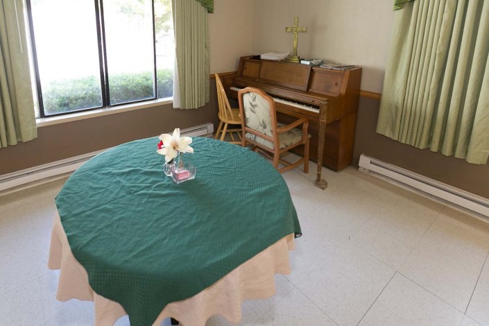 A room with a piano and chair in the corner, a round table with a green tablecloth, and a flower arrangement on top.