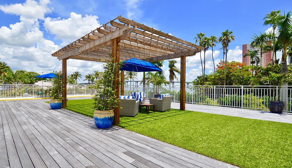 A sunny outdoor seating area with a pergola and umbrella on a wooden deck, surrounded by a garden and palm trees.