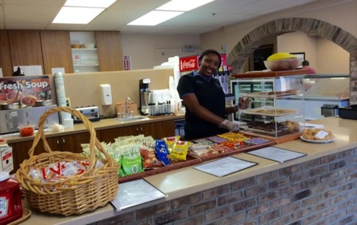 An employee stands behind a cafe counter displaying various snacks and beverages.
