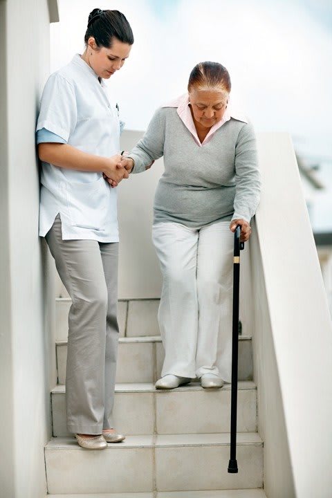 A caregiver assisting an elderly woman with a cane down the stairs.