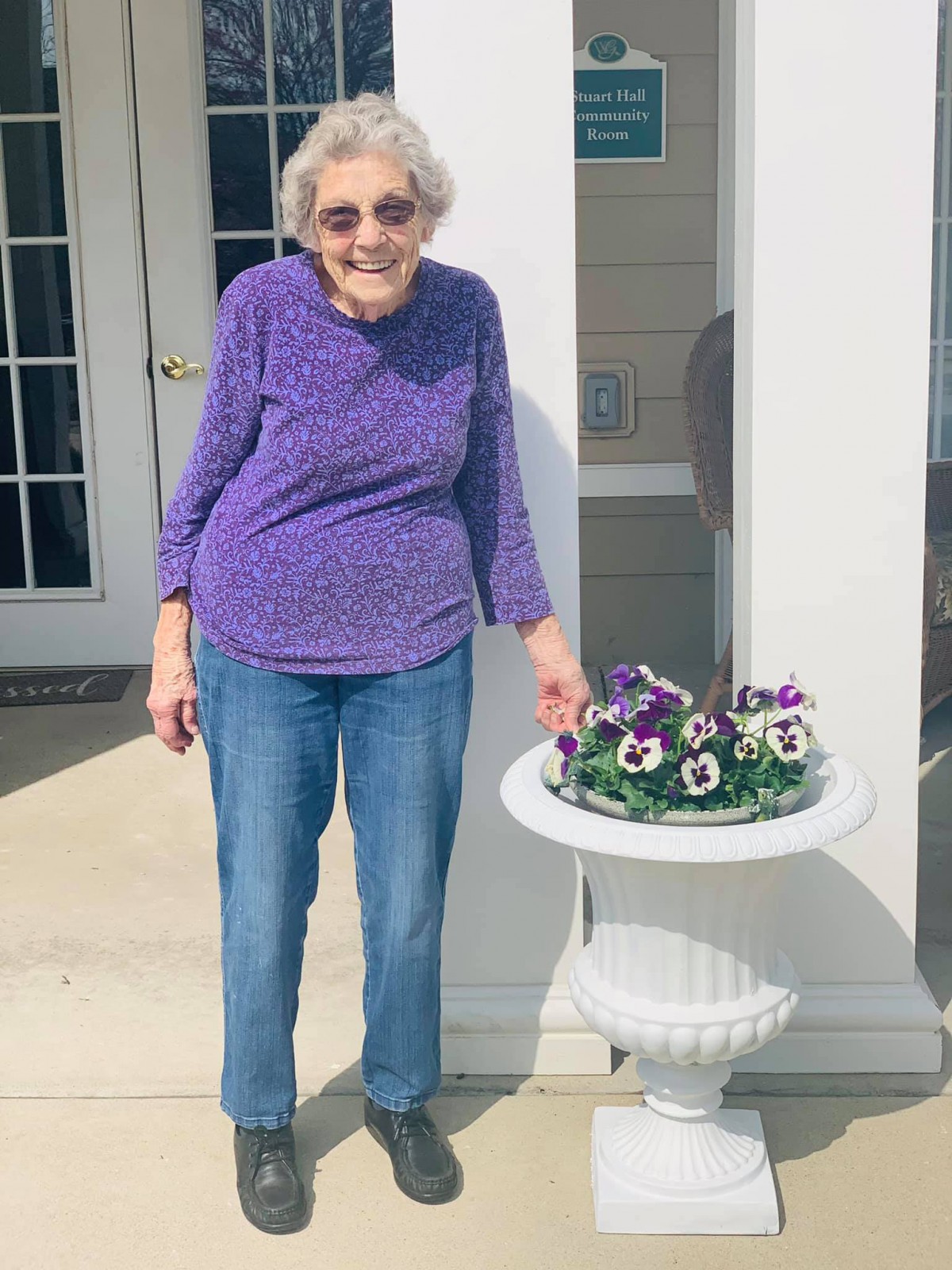 An elderly woman smiles as she stands next to a flower pot outdoors.