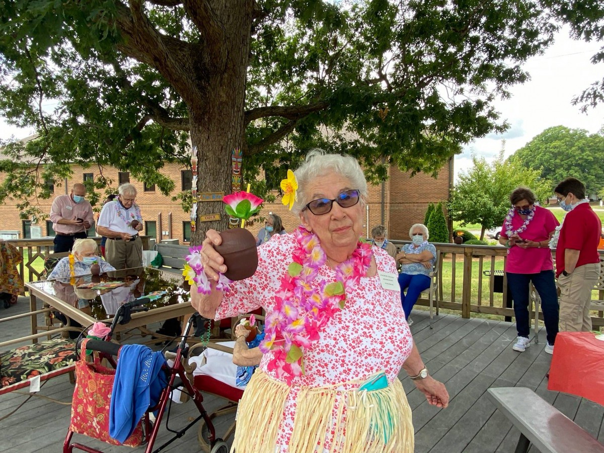 An elderly woman enjoys a Hawaiian-themed celebration outdoors, dressed in a colorful costume with leis and holding a coconut cup.
