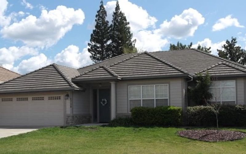 A single-story suburban house with a manicured front yard and trees in the background under a cloudy sky.