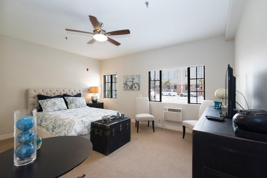 A modern bedroom interior with a bed, ceiling fan, and large windows.