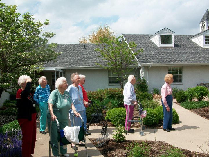 A group of elderly individuals walking outdoors in a garden setting at a retirement home.