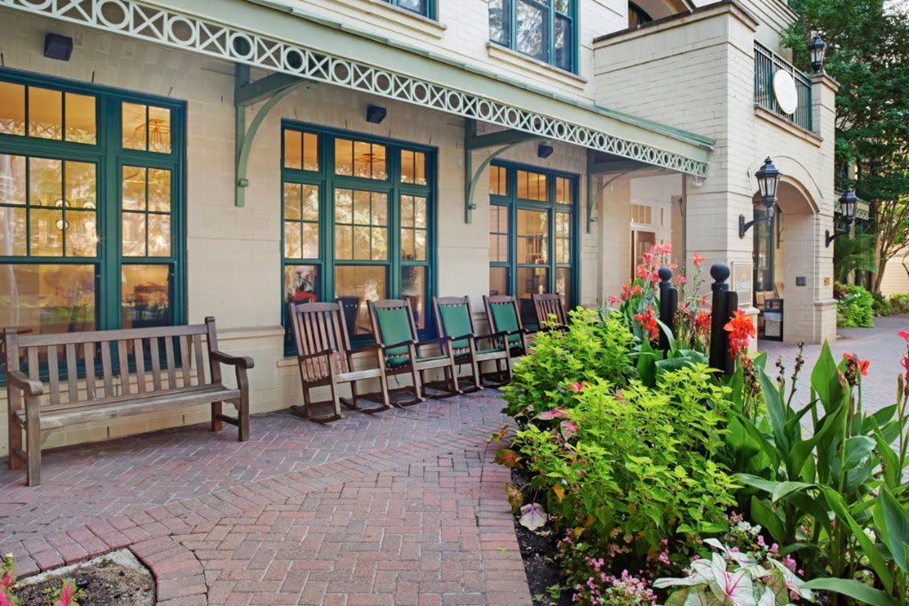 A cozy outdoor patio area with benches and rocking chairs, surrounded by lush greenery and a charming building facade.
