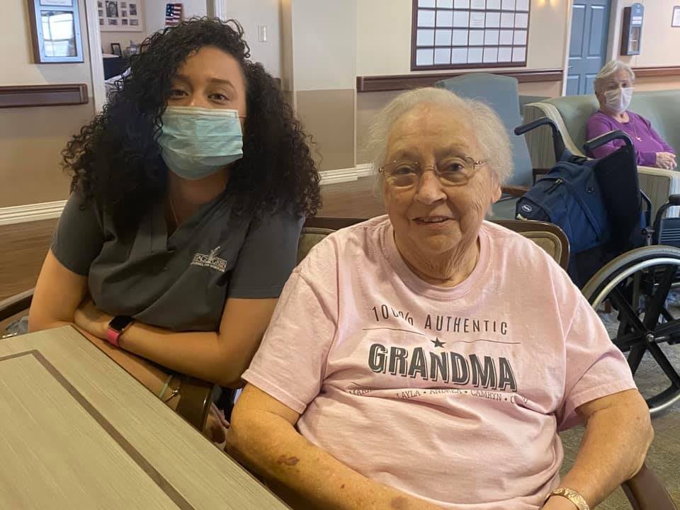 An elderly woman sitting next to a caregiver who is wearing a mask.