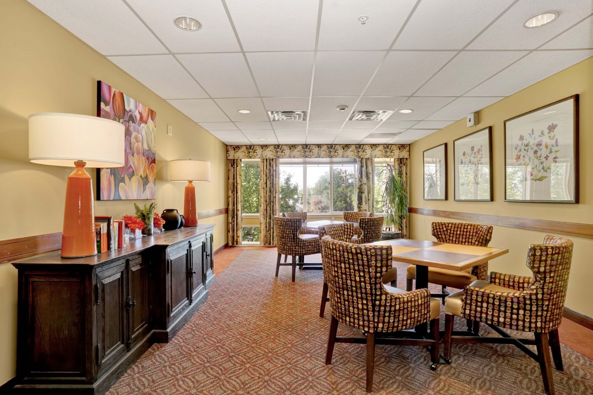 A cozy dining area with a table and chairs, decorated with artwork and lamps, offering a view through large windows.