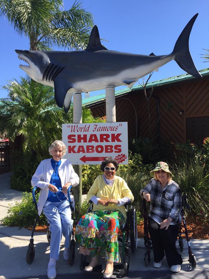 Three people pose in front of a 'World Famous Shark Kabobs' sign with a shark statue overhead.