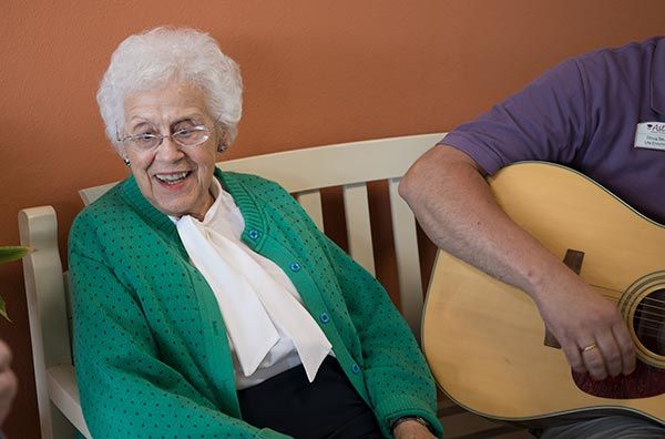 An elderly woman smiling while sitting next to someone playing the guitar.