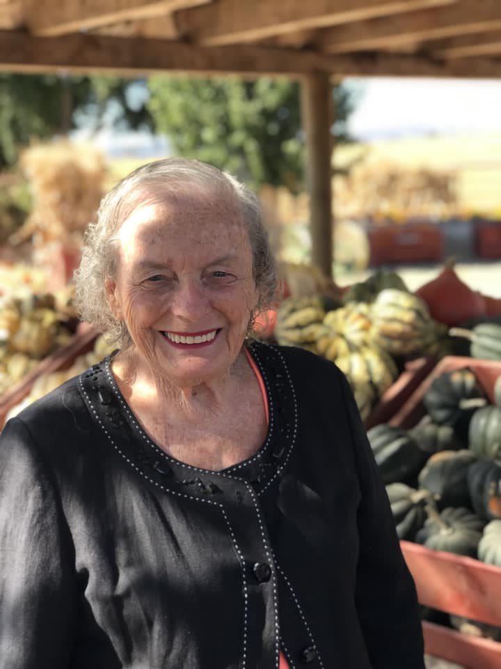 An elderly woman smiling in front of a display of vegetables outdoors, possibly during harvest season.