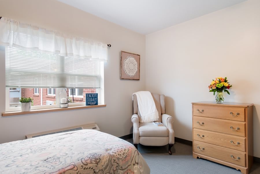 A cozy bedroom with a floral bedspread, an armchair by the window, and a wooden dresser topped with flowers.