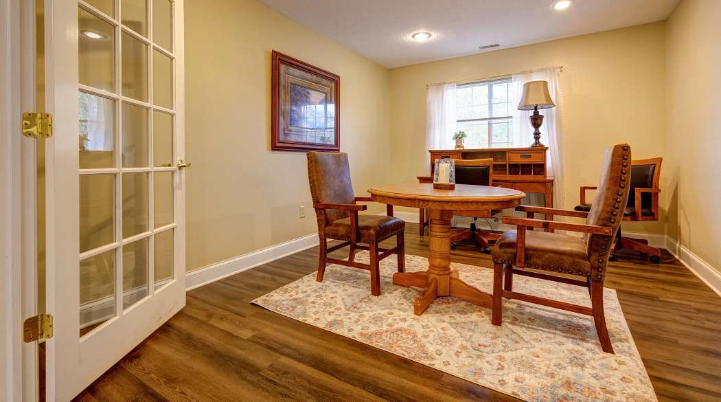 A cozy home office with a wooden desk and chairs, featuring a rug and a lamp by the window.