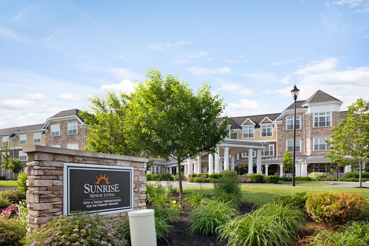 A large residential building surrounded by a well-maintained garden with a tree and a prominent sign in the foreground.