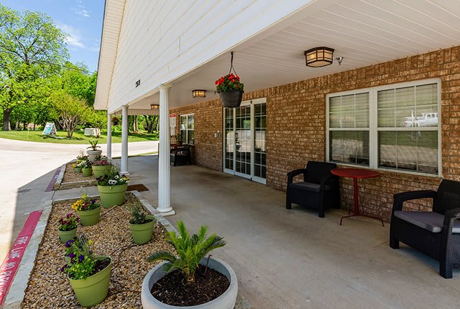 A covered porch with brick walls, featuring potted plants, patio furniture, and decorative columns.