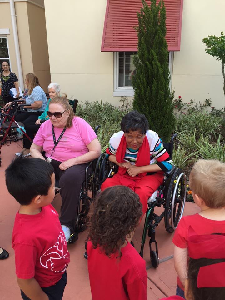 Children interacting with elderly individuals in wheelchairs outside a care facility.