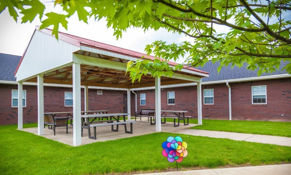 An outdoor pavilion with picnic tables surrounded by a grassy lawn and trees.