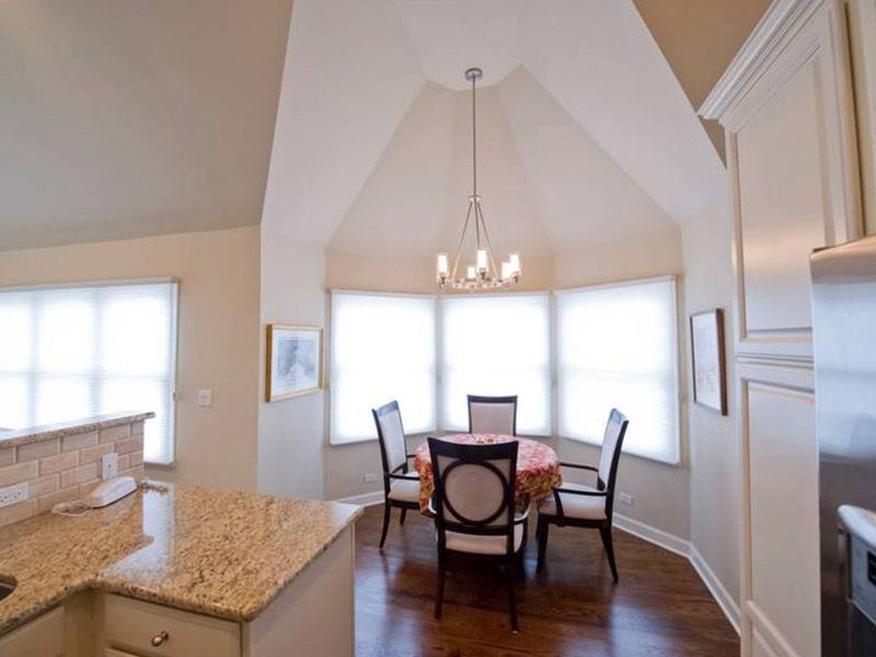 A cozy dining area with a round table, elegant chairs, and a chandelier, illuminated by abundant natural light from large windows.