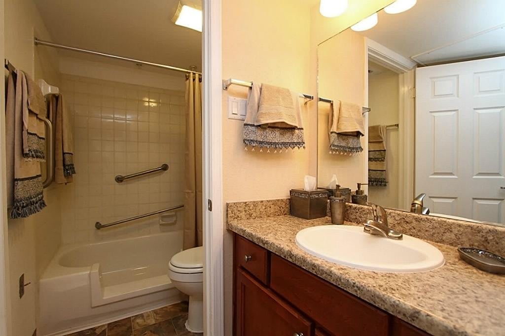 A well-lit bathroom featuring a granite countertop sink, mirror, and a shower with safety bars.