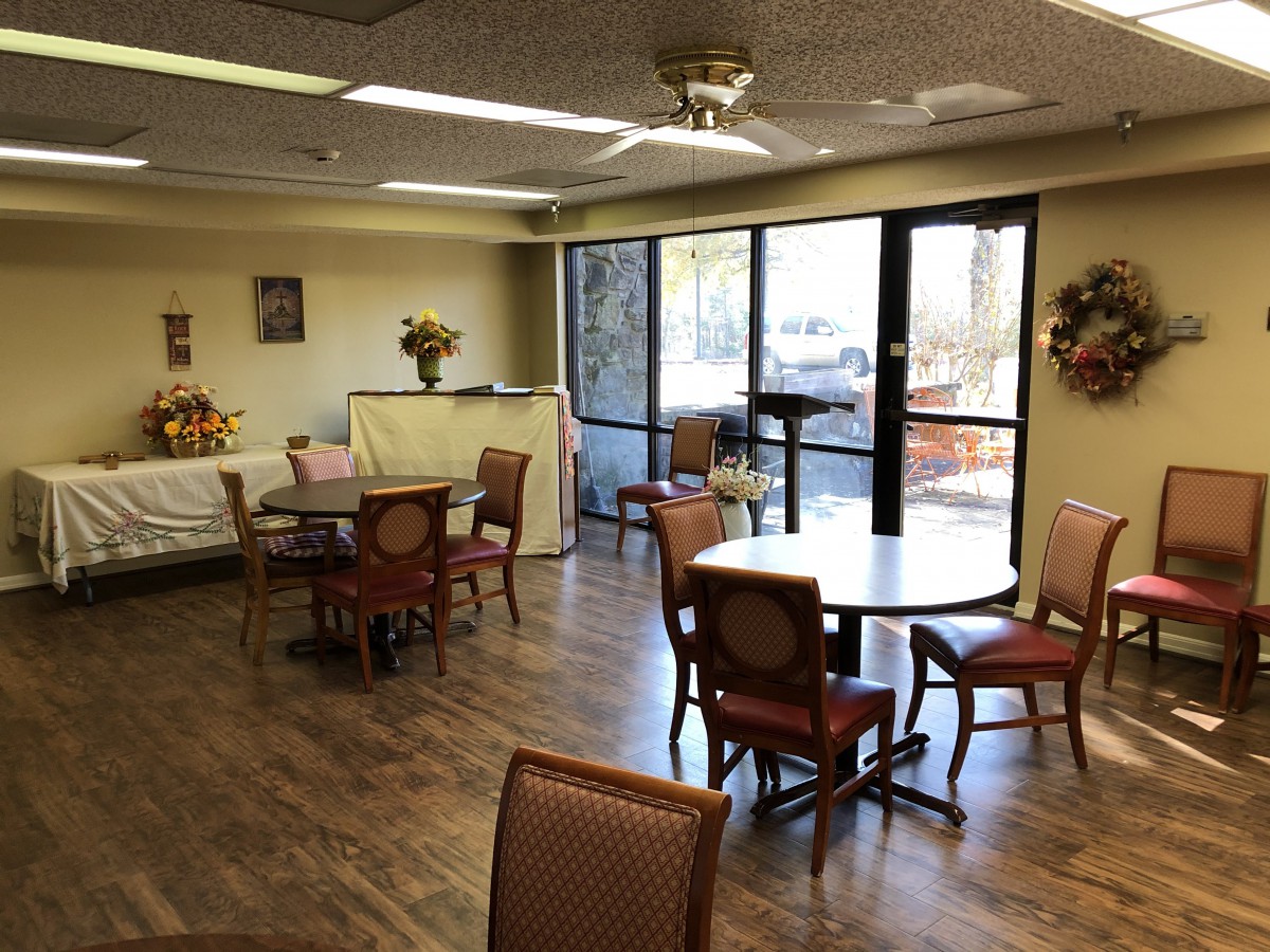 A cozy dining area with round tables and upholstered chairs, featuring large windows and a ceiling fan.