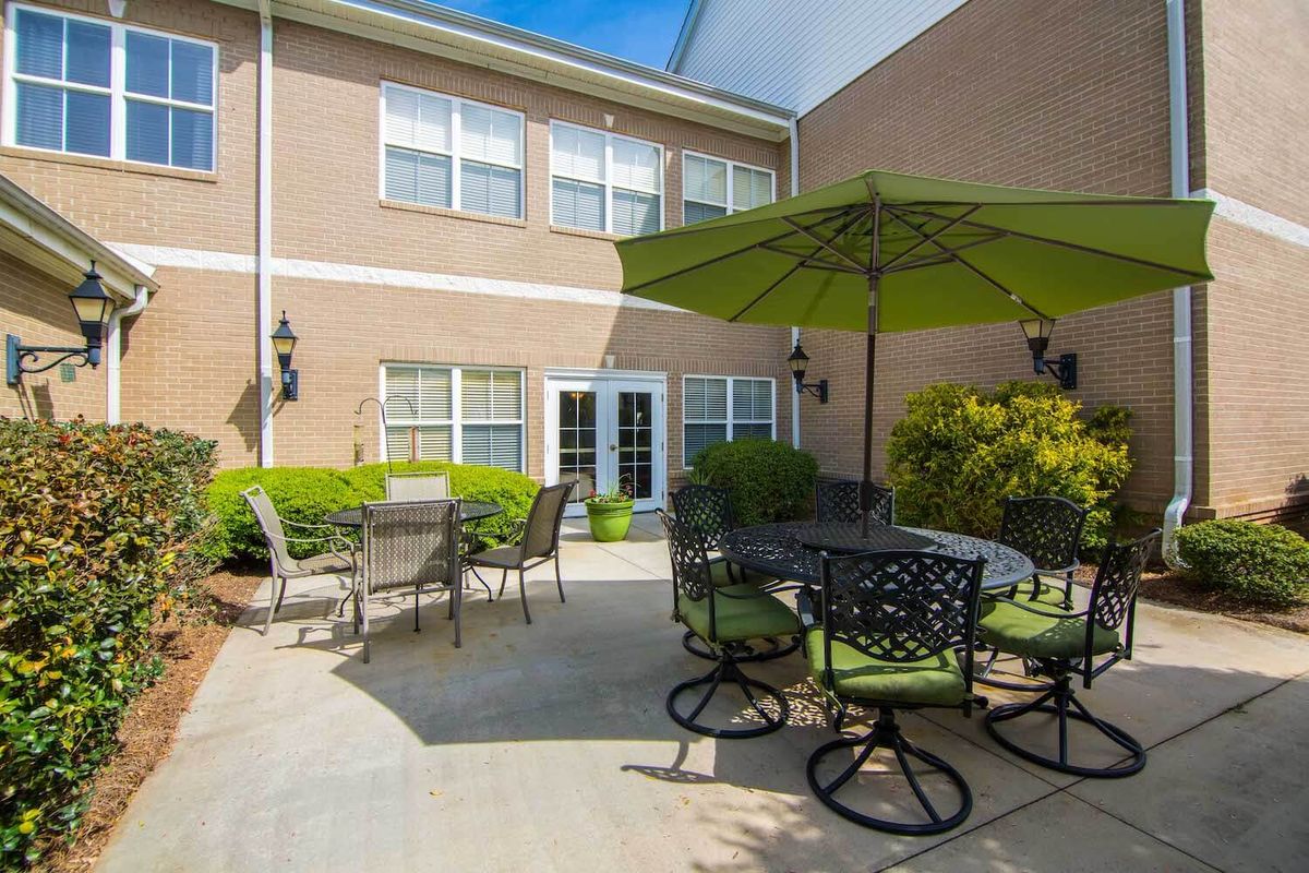 A cozy outdoor patio with tables, chairs, and a large umbrella, surrounded by greenery on a sunny day.