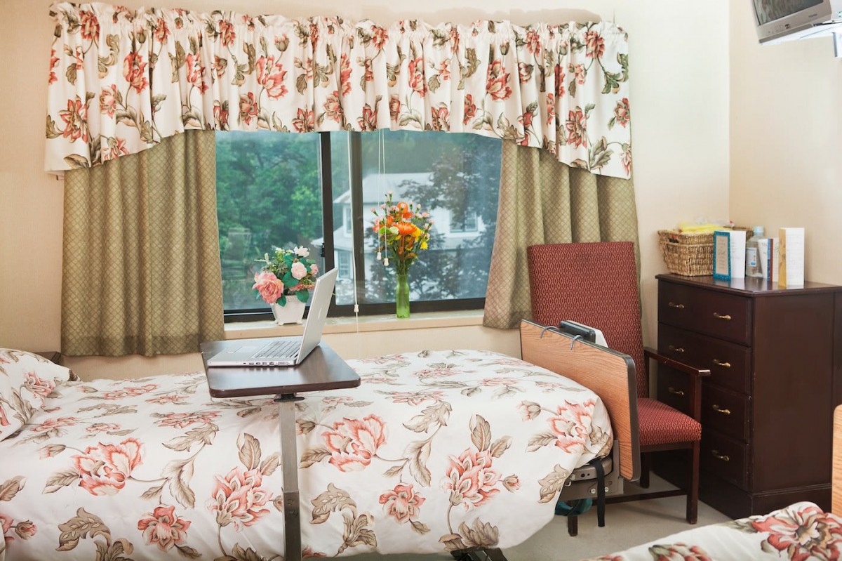 A cozy bedroom with floral decor, featuring a laptop on the bed, and a scenic window view.