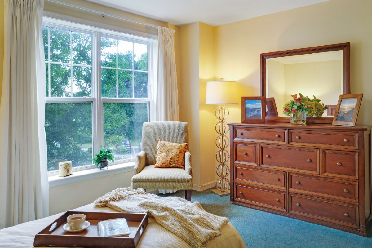 A cozy bedroom featuring a large window, comfortable armchair, and wooden dresser with a mirror.
