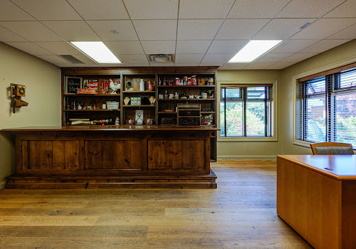 A modern office interior featuring a wooden reception desk, shelves filled with various items, and large windows allowing natural light.