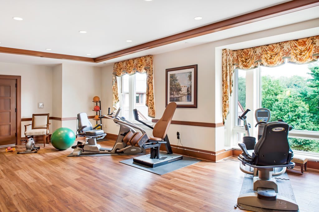 A well-lit home gym with various exercise equipment on wood flooring.