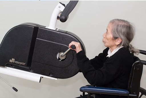 An elderly person using a rehabilitation exercise machine while seated in a wheelchair.