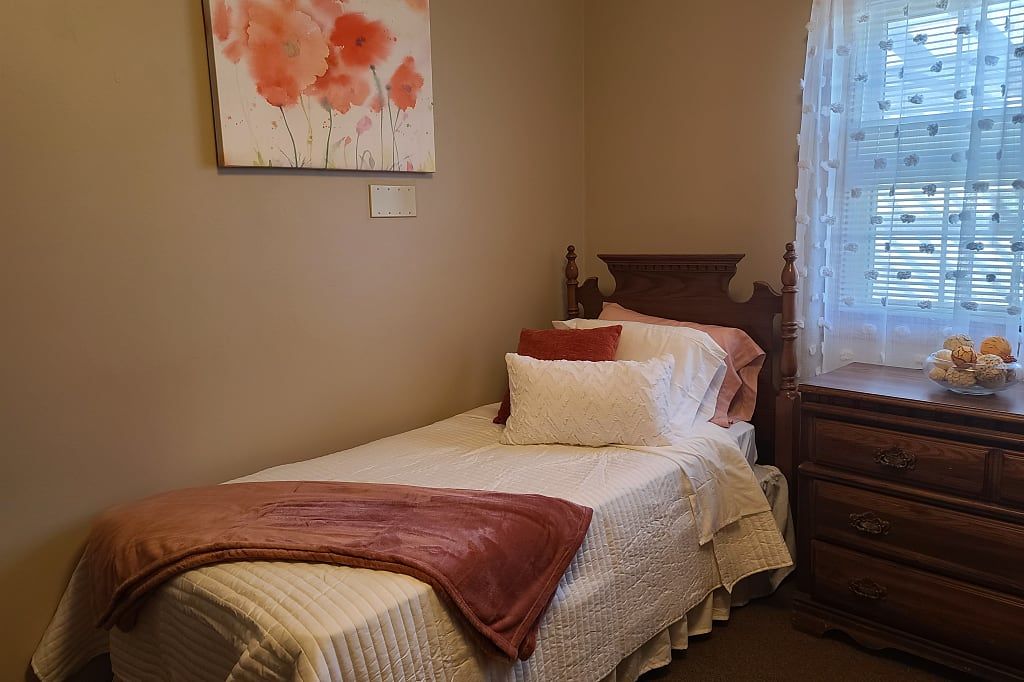 A cozy bedroom with a single bed, adorned with pillows and a blanket, featuring artwork on the wall and natural light from a window.