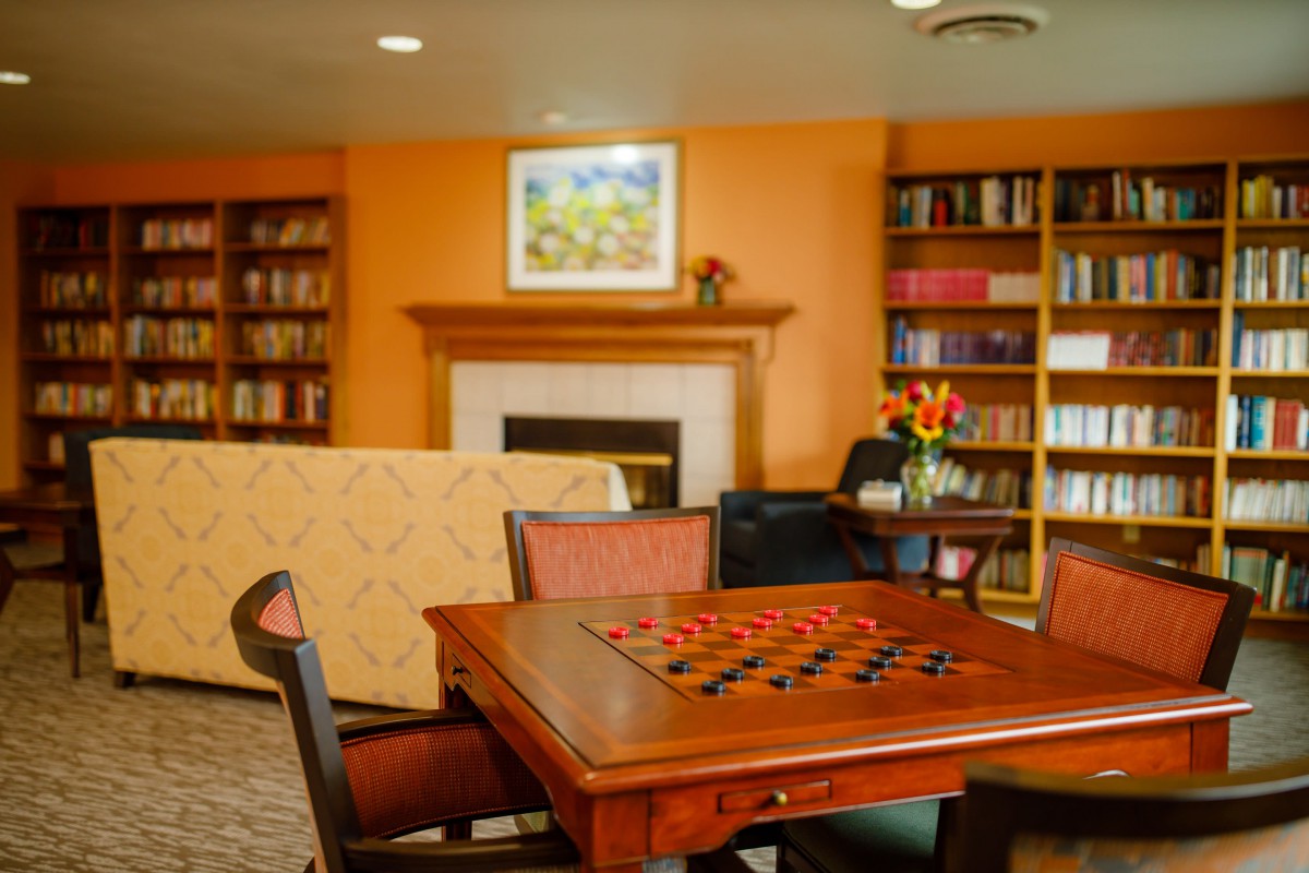 A cozy living room with a game of checkers set up on the table, surrounded by bookshelves and a fireplace.
