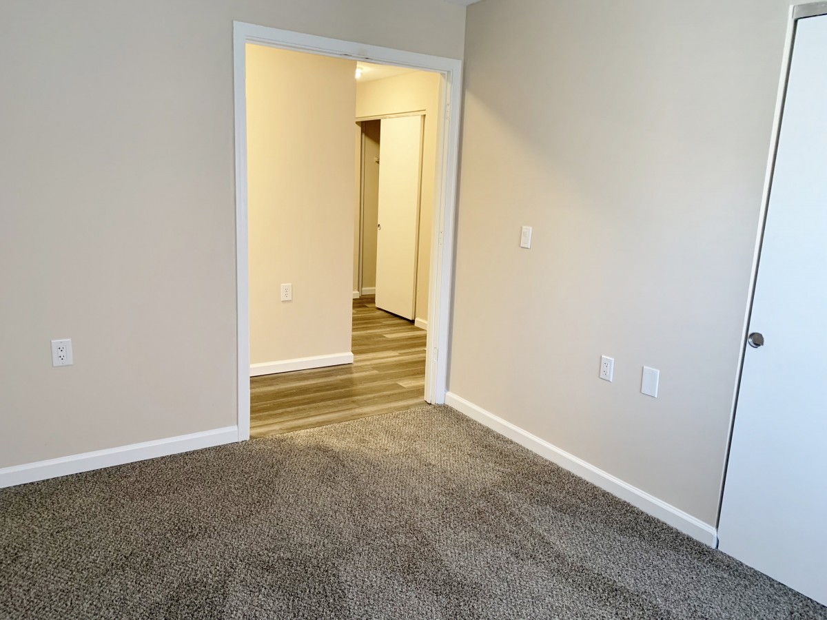 An image of an empty room with carpet flooring, leading to a hallway through a doorway.