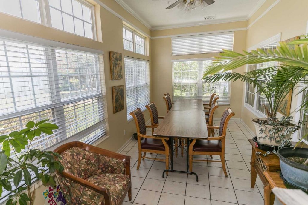 A well-lit dining area with large windows, featuring a long wooden table surrounded by chairs and adorned with indoor plants.