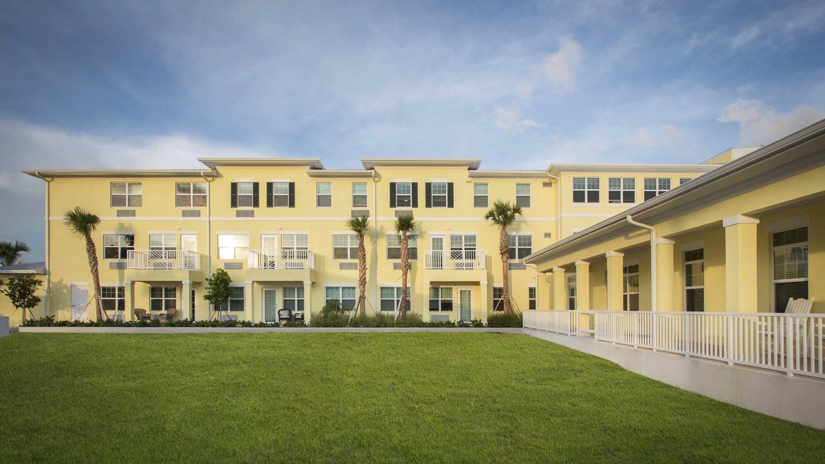 A yellow residential building with multiple floors, surrounded by palm trees and a grassy lawn.