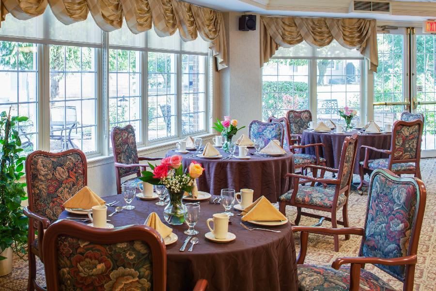 A cozy dining area with elegantly set tables and chairs, adorned with floral centerpieces near large windows.