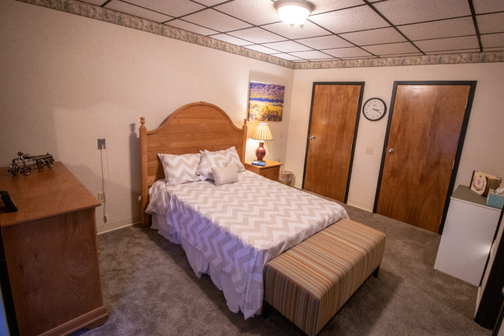 A cozy bedroom with a wooden bed frame, patterned bedding, and a striped bench at the foot of the bed. The room includes a lamp on a nightstand and two wooden doors.
