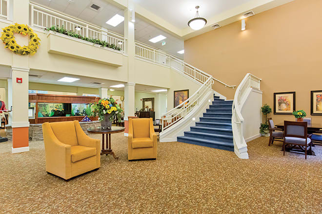 A spacious lobby featuring a carpeted floor, comfortable seating arrangements, a prominent staircase, and decorative flowers.