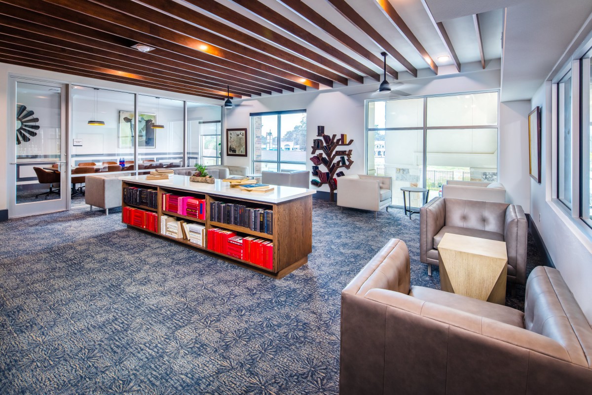 A modern office space with open layout, featuring wooden ceiling beams, stylish furniture, bookshelves, and ample natural light.