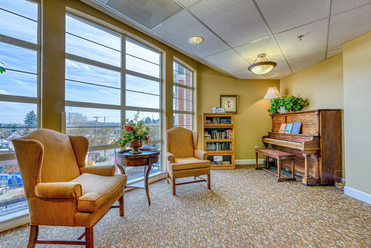 A cozy lounge with a piano, chairs, and a bookshelf by a large window.