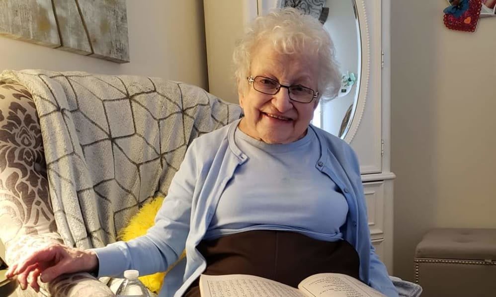 An elderly woman smiles while sitting comfortably in a chair with an open book.