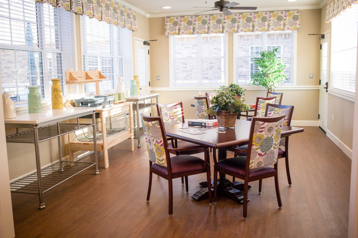 A well-lit dining room with a round table surrounded by chairs, featuring large windows and a hardwood floor.
