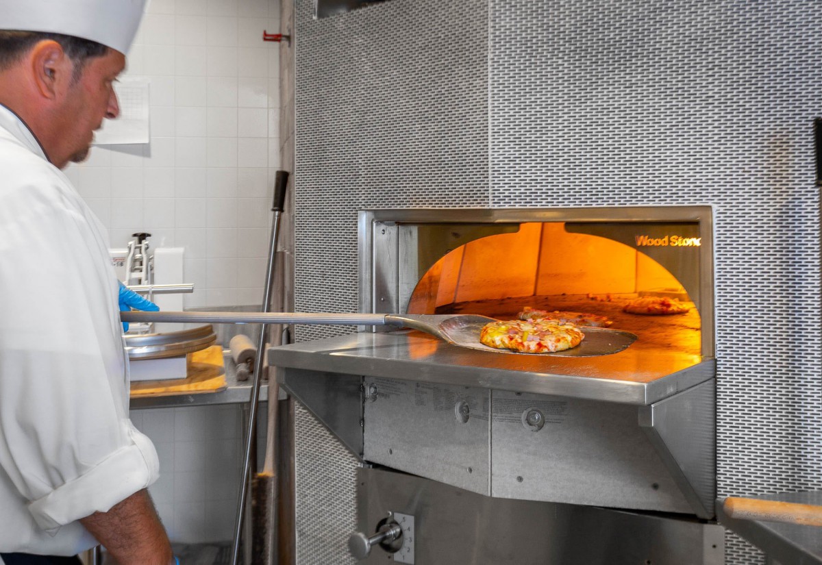 A pizza being placed into a wood-fired oven in a restaurant kitchen.