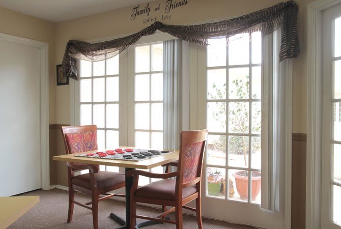 A cozy dining area featuring a small table and two chairs, illuminated by daylight streaming through large windows with elegant curtains.